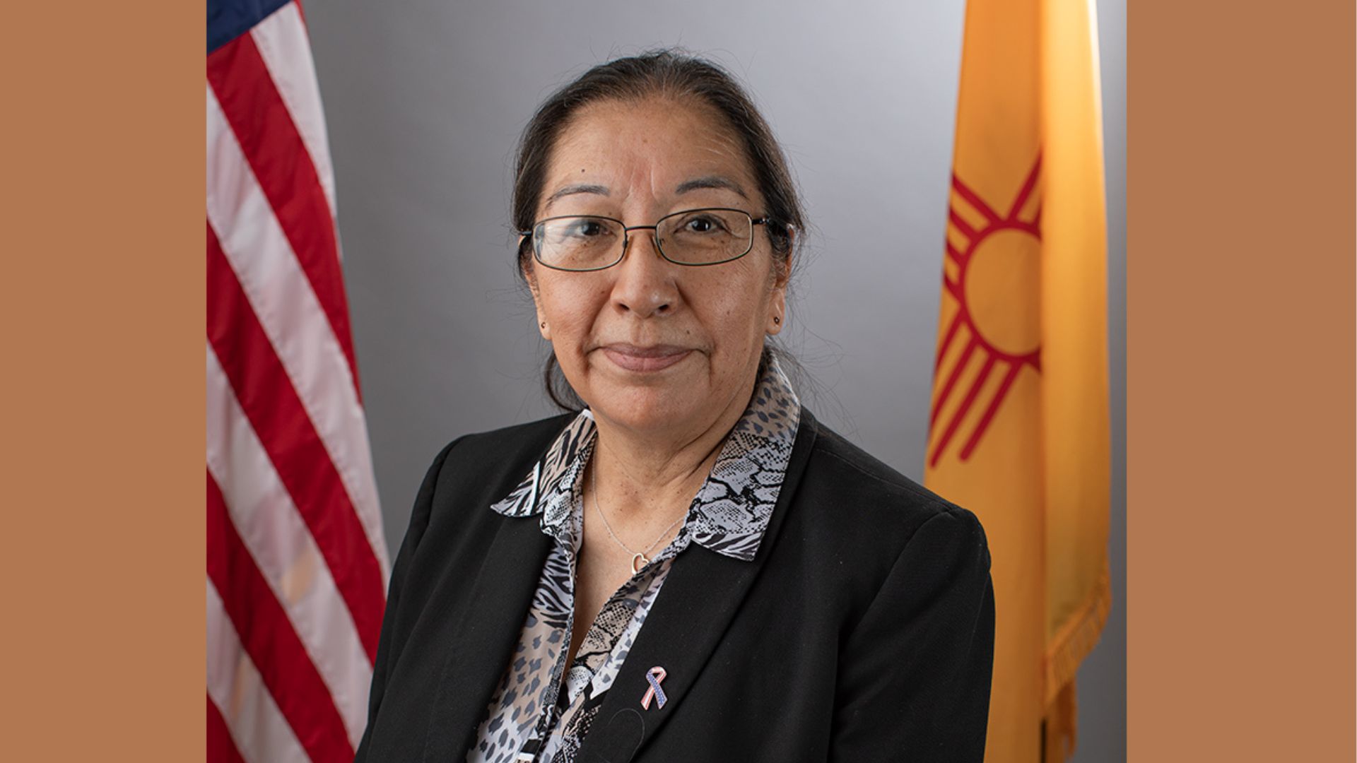 A woman wearing glasses and a black blazer stands in front of the U.S. flag and the New Mexico state flag.