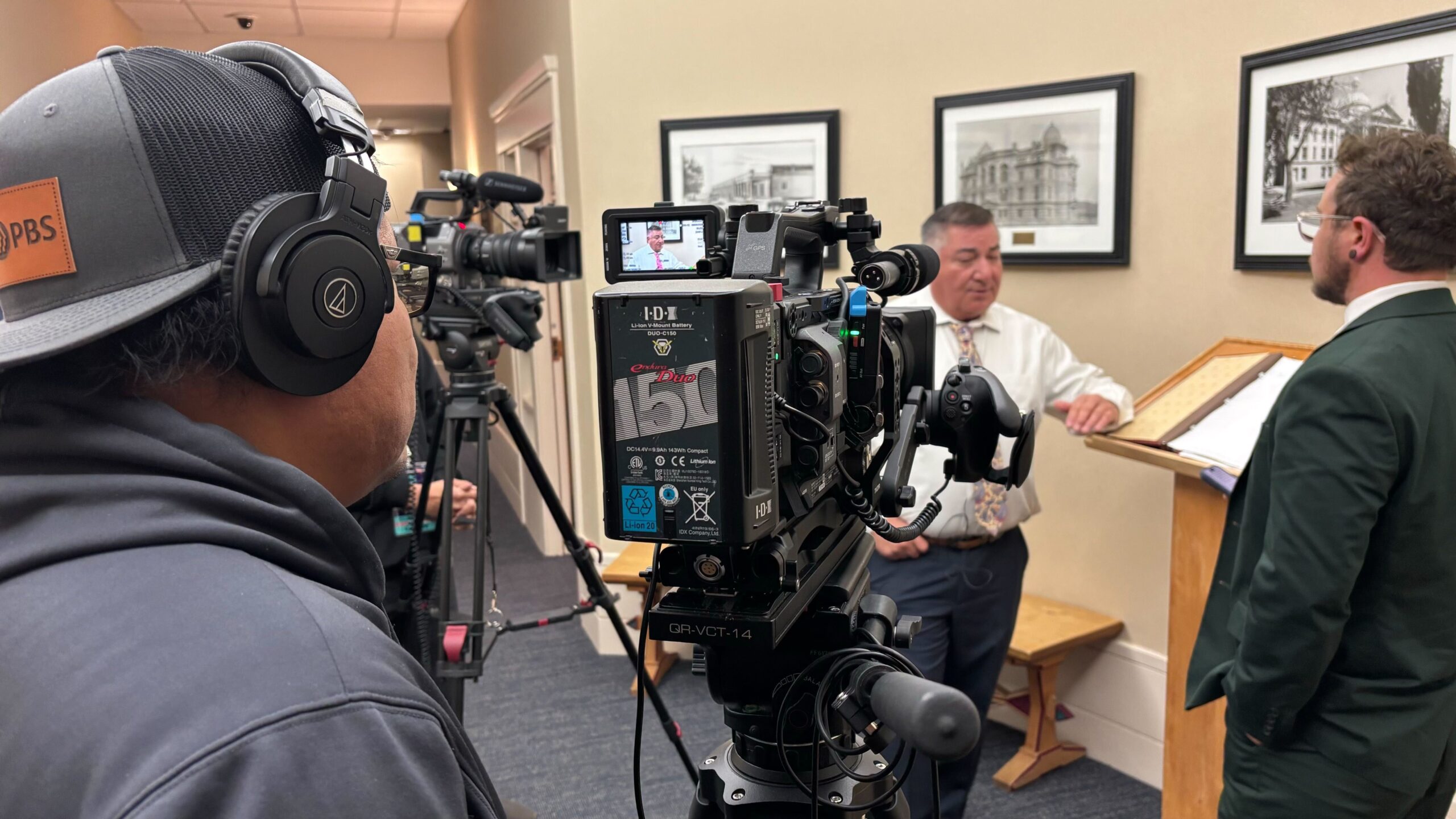A camera crew films two men in business attire having a conversation in an office hallway with framed pictures on the wall.