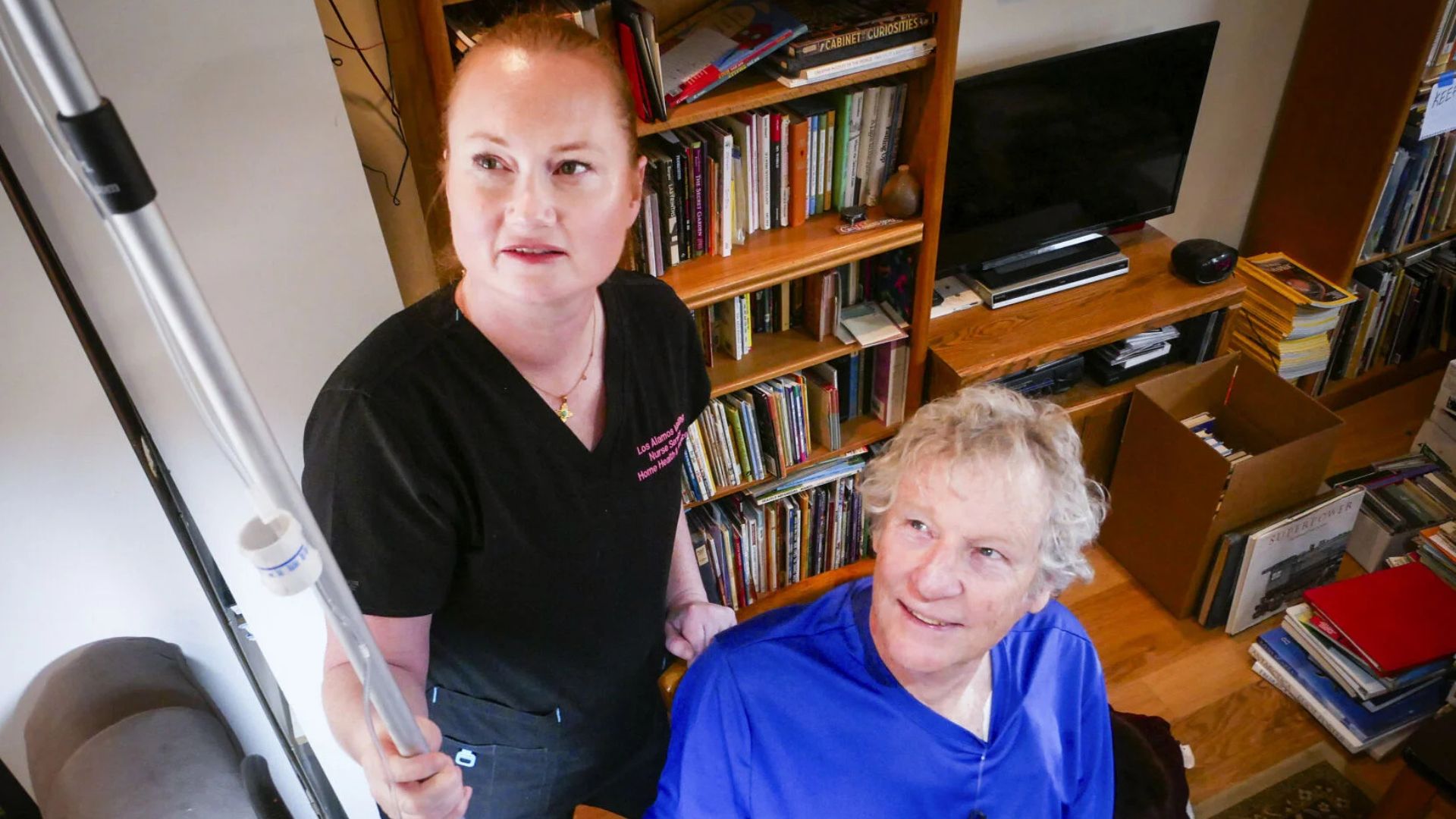A woman in scrubs holds a pole-like device while standing next to an older man seated in a room filled with bookshelves and assorted items.