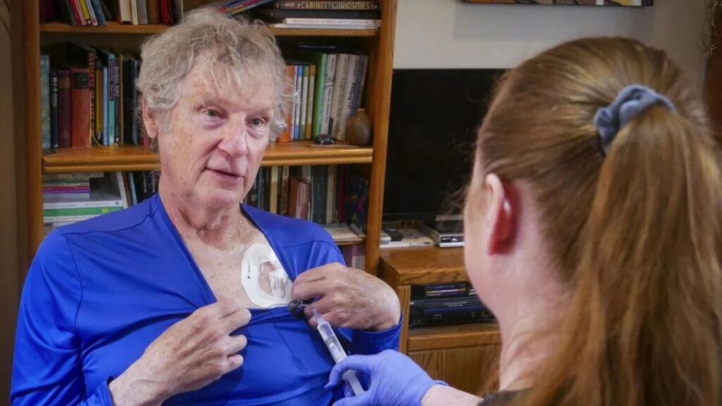 An older man pulls down his shirt to reveal a port on his chest while a woman wearing gloves attends to it with a syringe.