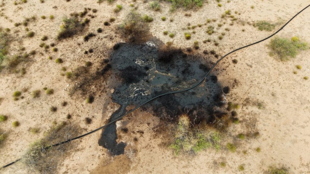 Aerial view of an oil spill on dry, sandy terrain with sparse vegetation and a black hose or pipe running across the scene.