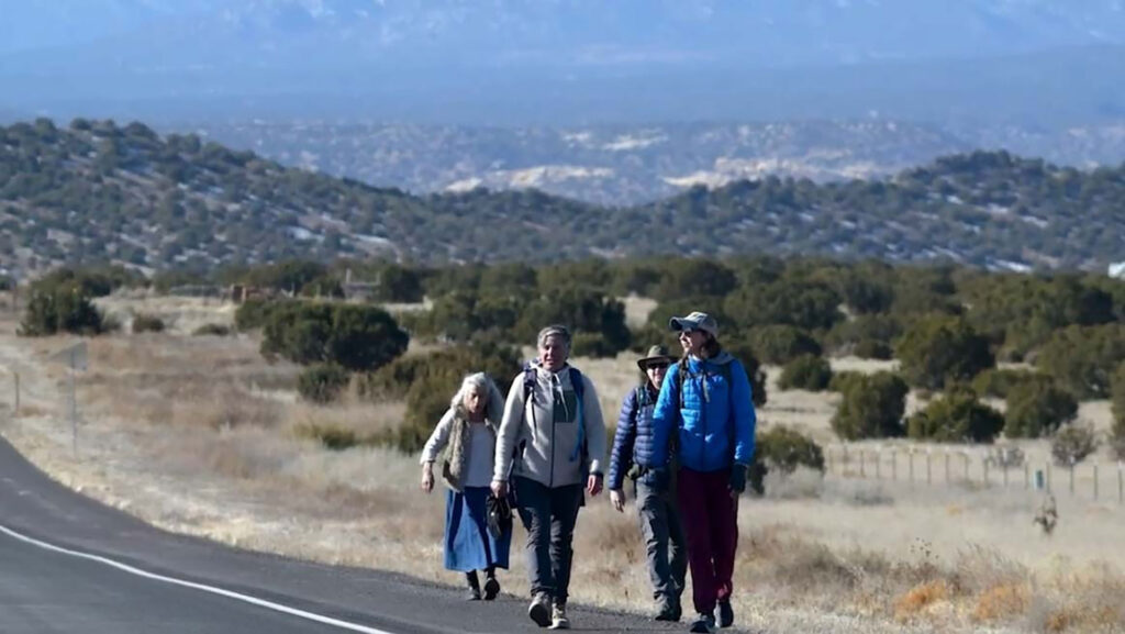 Four people walk along a rural road with sparse vegetation and distant hills visible under a clear sky.