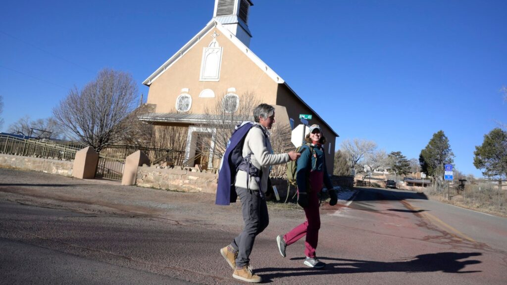 Two people with backpacks walk across a street in front of a tan church with a white steeple under a clear blue sky.