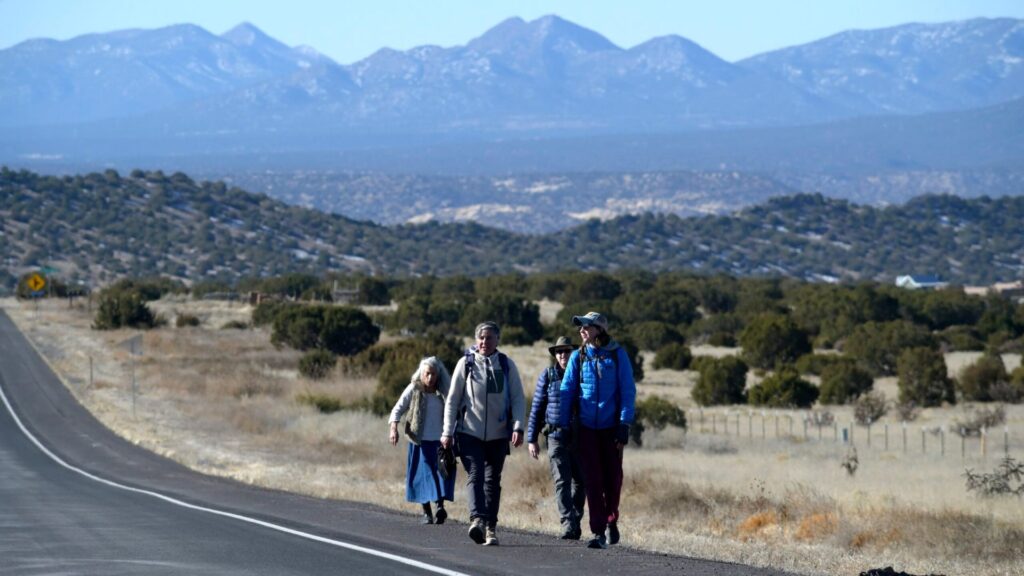 Four people walk along the side of a rural road with mountains and shrubs in the background under a clear sky.