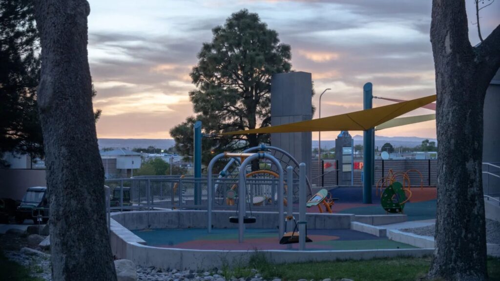 A playground with climbing structures and shade sails, surrounded by trees, is empty at sunset with buildings visible in the background.