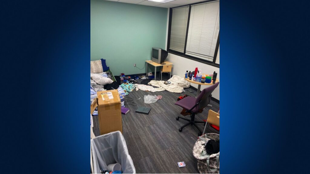 A cluttered room with a mattress on the floor, scattered belongings, a TV, cardboard boxes, a chair, trash bin, and laundry basket under fluorescent lighting.