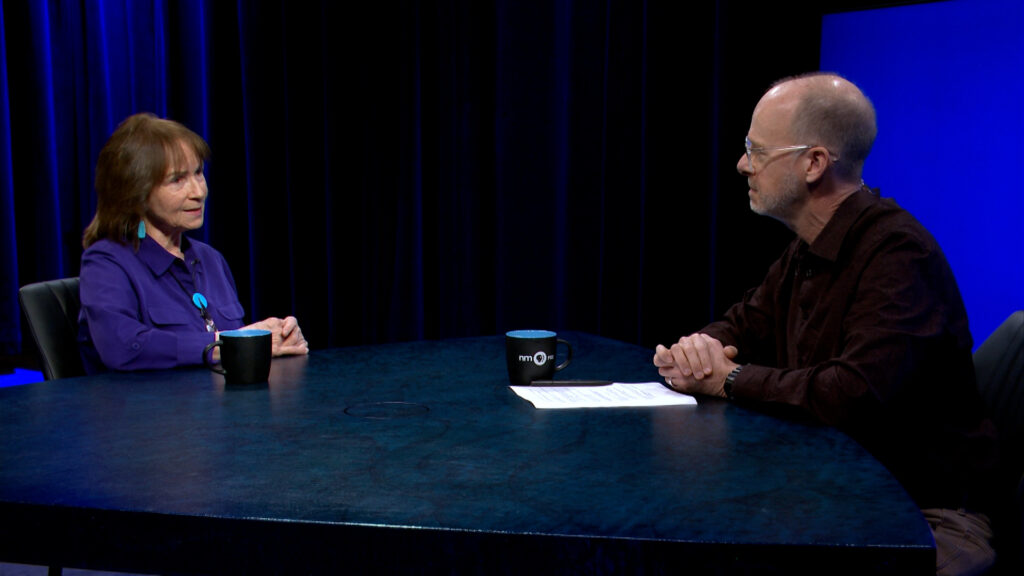 Two people sit across from each other at a round table with coffee mugs and papers, engaged in conversation in a dimly lit studio setting.