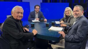 Four people sit around a table with notebooks, pens, and coffee mugs in a studio setting with blue screens in the background.