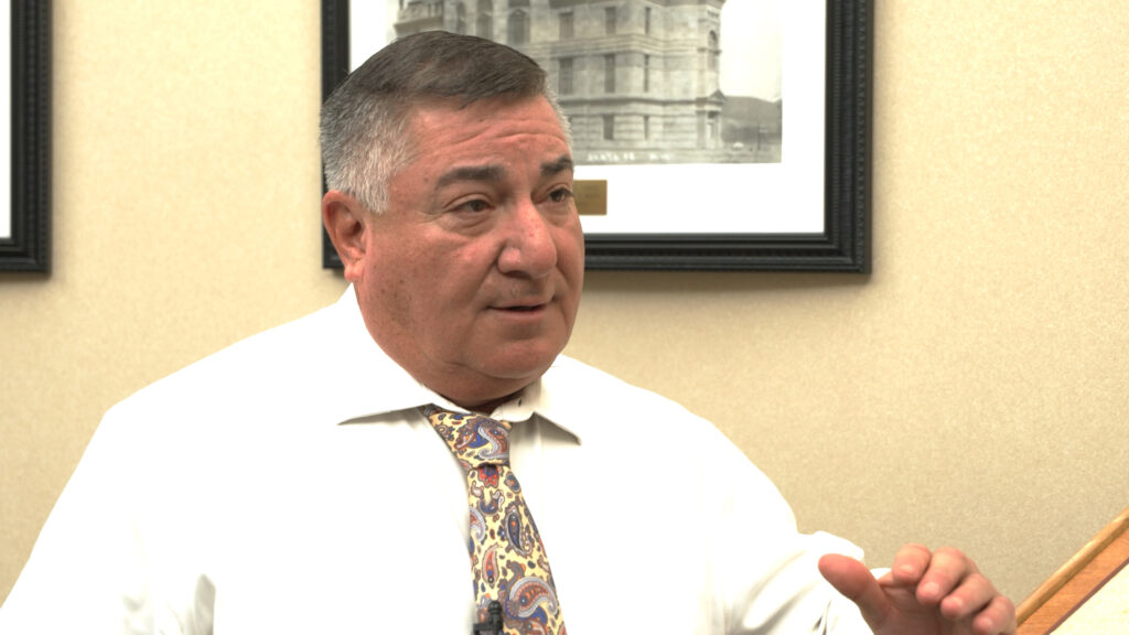 A man in a white shirt and patterned tie speaks, gesturing with his hand, with framed photos of buildings in the background.