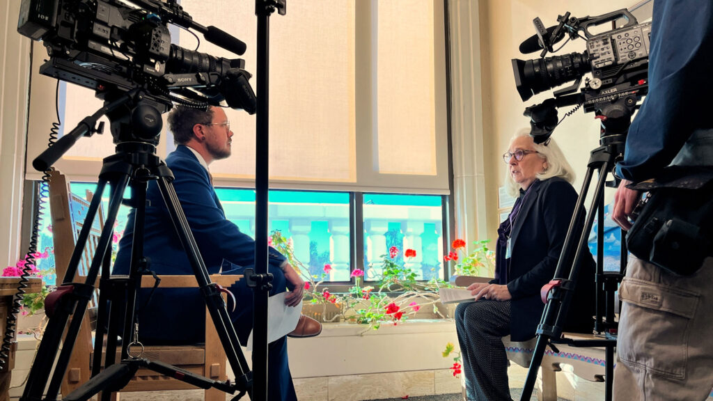 Two people sit facing each other for an interview, surrounded by cameras and lighting equipment, with flowers and large windows in the background.