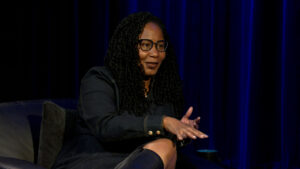 A woman with long curly hair and glasses speaks while seated on a stage against a dark blue curtain.