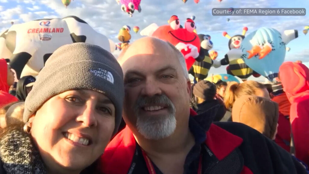 Two people smiling at a hot air balloon festival with various cartoon-themed balloons and a crowd in the background.