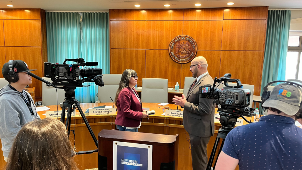 Two people speak at a city council meeting room while camera crews film them. Nameplates and a city seal are visible on the wooden wall behind them.