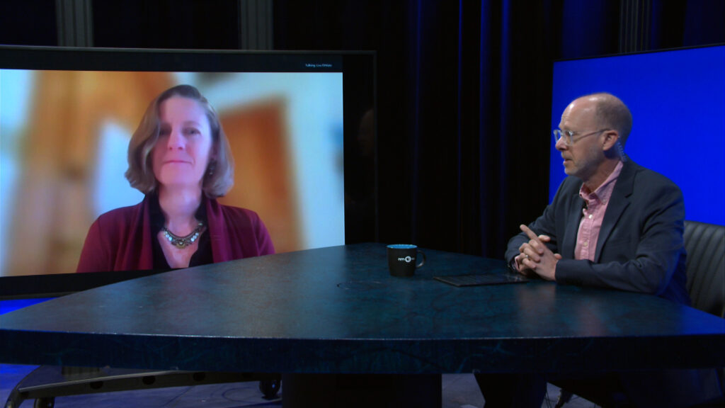 A woman appears on a large video screen while a man sits at a table, facing the screen, in a dimly lit studio setting.
