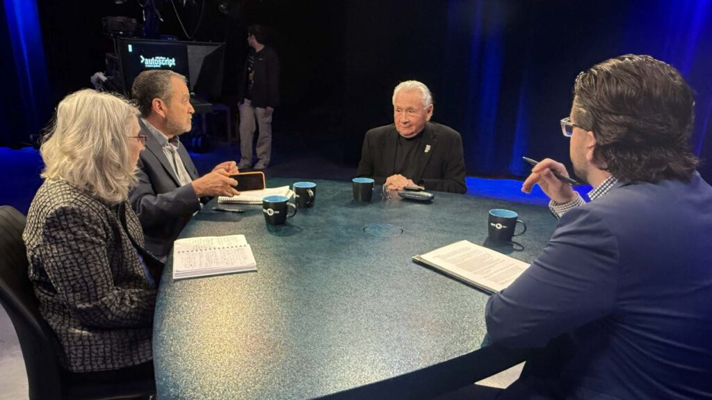 Four people sit around a round table with notebooks, mugs, and papers, engaged in discussion in a studio setting with cameras and lighting equipment in the background.