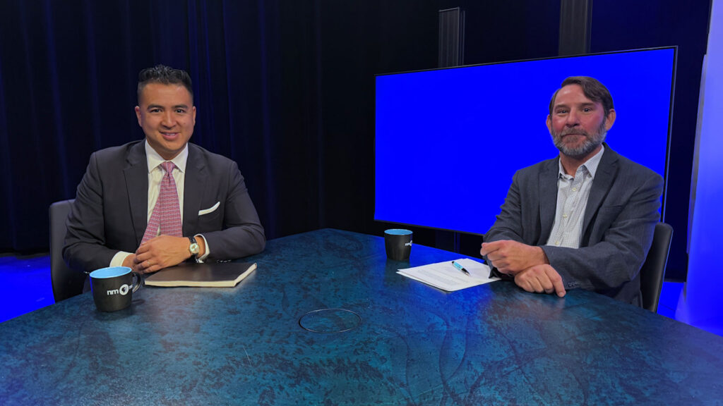 Two men in suits sit at a table in a studio setting with a blue screen in the background, each with a mug and papers in front of them.