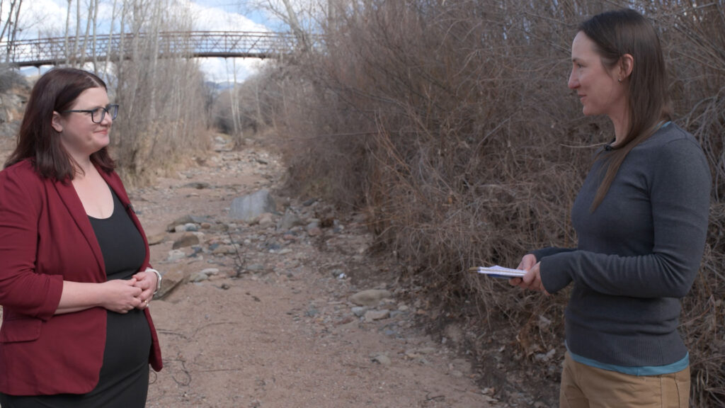 Two women stand and talk in a dry, rocky creek bed with leafless bushes and a pedestrian bridge in the background.