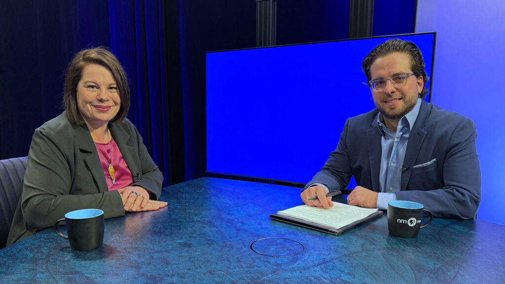Two people sit at a table in a studio with a blue screen in the background. They both have mugs, and one holds a notepad and pen.