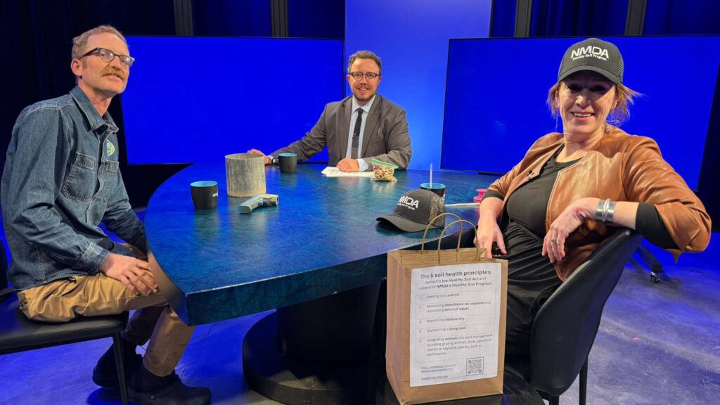 Three people sit around a blue studio table with mugs, papers, and a bag labeled "10 soil health principles." Two wear ANIDA hats, and studio lights are visible in the background.