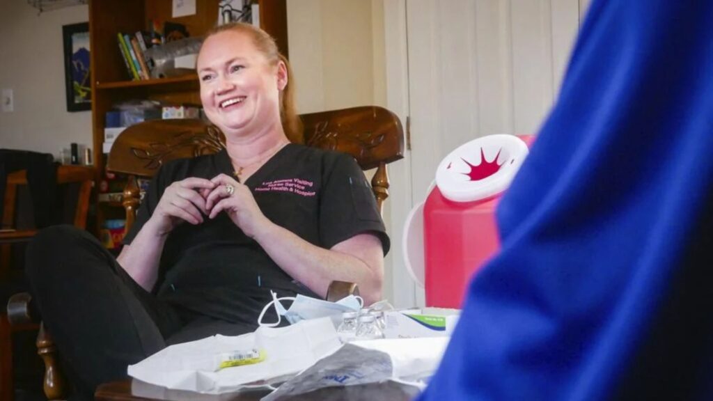 A woman in black scrubs sits and smiles in a room with medical supplies on the table in front of her and a red biohazard container nearby.