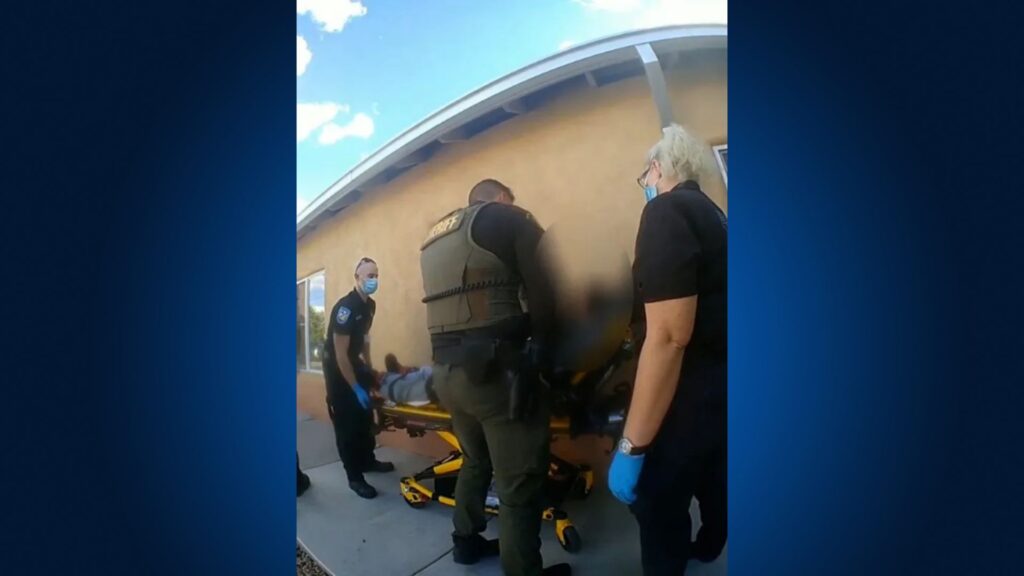 Several emergency responders and a law enforcement officer assist a person on a stretcher outside a building.