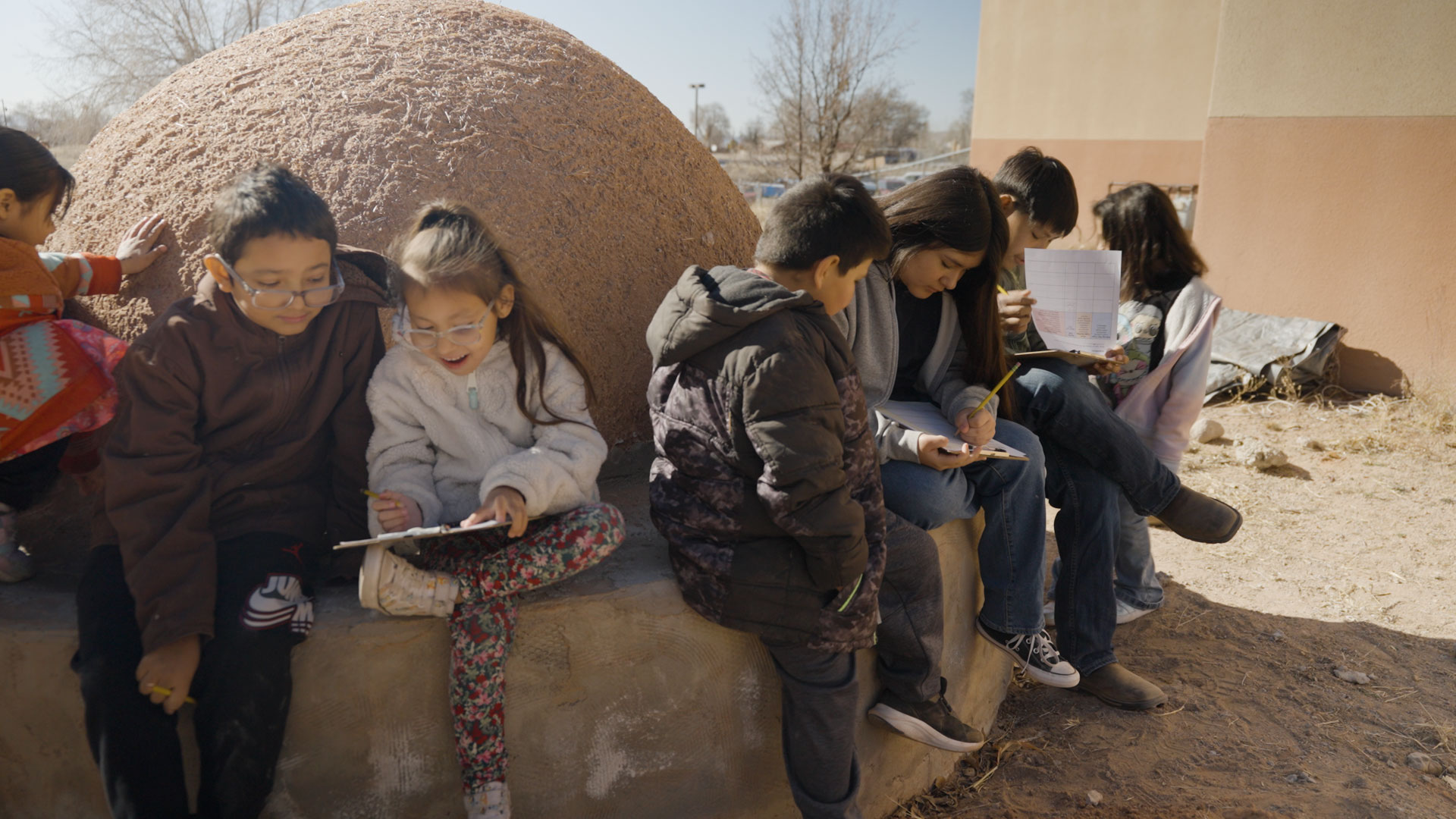 A group of children sit and stand outside by a rounded structure, writing or drawing on paper, with a building and leafless trees in the background.