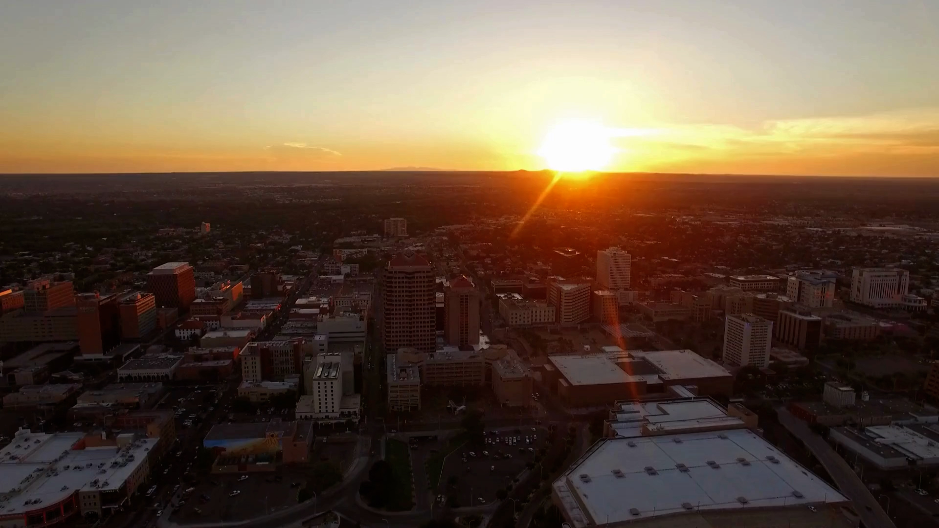 Aerial view of a city skyline at sunset, with the sun low on the horizon casting a warm glow over buildings and streets.