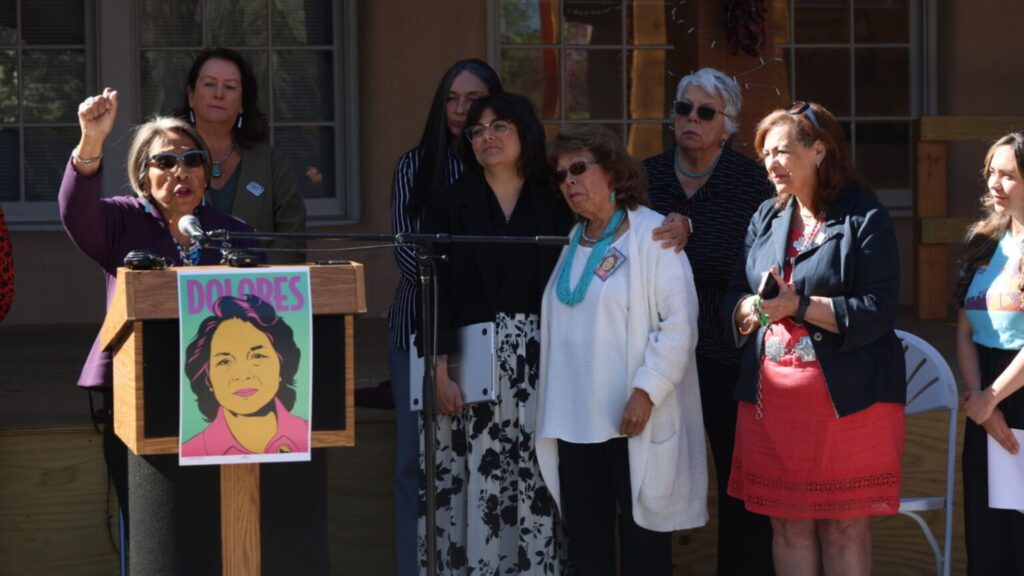 A group of women stand together at an outdoor event. One woman speaks at a podium with a "Dolores" poster, while others listen attentively.
