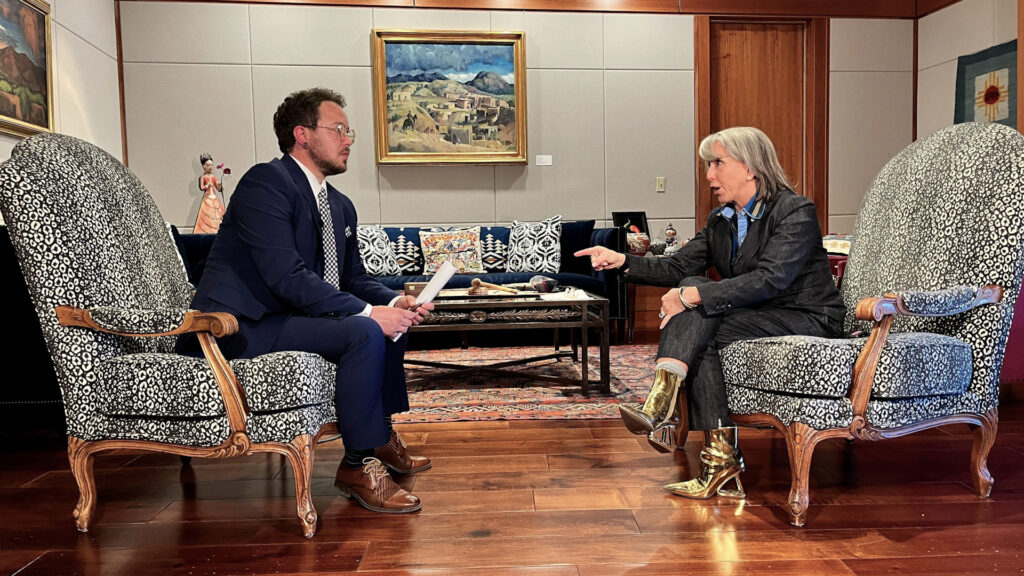 Two people sit facing each other in armchairs, engaged in conversation in a formal, art-filled office or living room. One holds papers; the other gestures while speaking.
