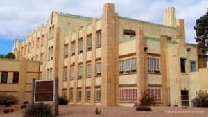 A beige, three-story Art Deco building with vertical lines and barred windows; a brown sign stands in front on a dry, landscaped area.