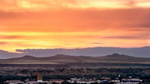 A wide view of a town with low buildings in the foreground and hills in the background under an orange and yellow sunset sky.