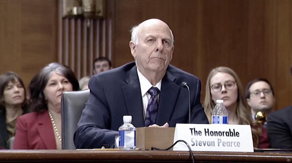 An older man in a suit sits at a desk with a nameplate reading "The Honorable Stevan Pearce" during a formal hearing, with several people seated in the background.