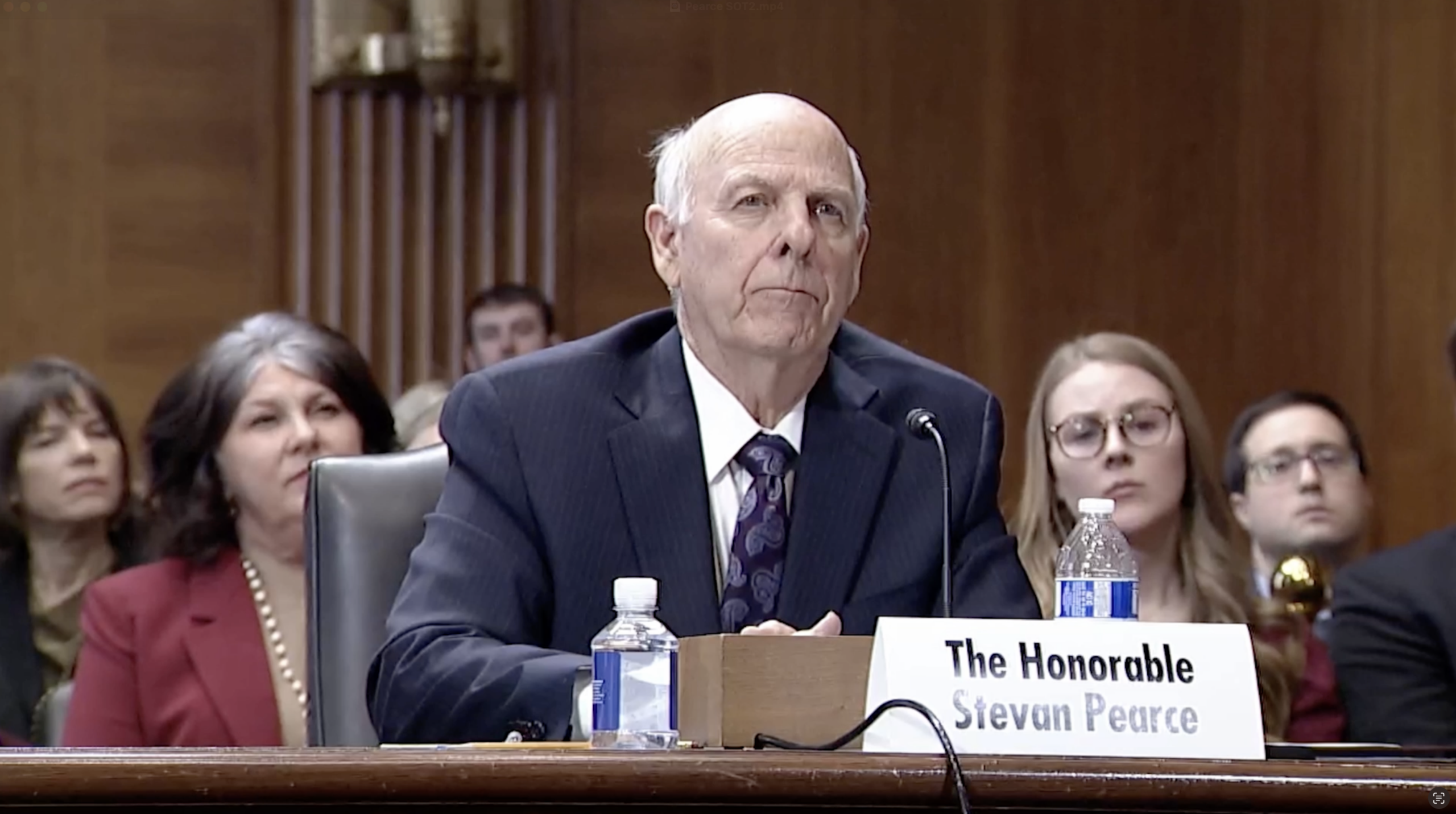 An older man in a suit sits at a hearing table with a nameplate reading "The Honorable Stevan Pearce." Several people sit in the background.