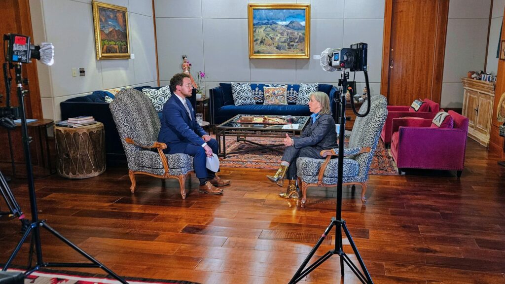 Two people are seated across from each other in armchairs, having a formal conversation in a well-lit, art-filled living room set up for an interview with cameras and lights.