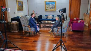 Two people are seated across from each other in armchairs, having a formal conversation in a well-lit, art-filled living room set up for an interview with cameras and lights.