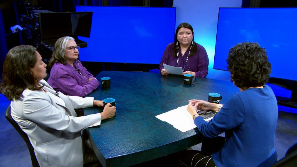 Four women sit around a round table in a TV studio, engaged in discussion. Three hold mugs, and one holds papers, with large screens and studio lights in the background.