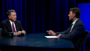 Two men in suits sit across from each other at a table in a studio with dark blue lighting; one holds a pen and paper, appearing to interview the other.