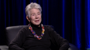 An older woman with short gray hair, wearing a black turtleneck and colorful necklace, sits in a black chair against a dark blue curtain background.