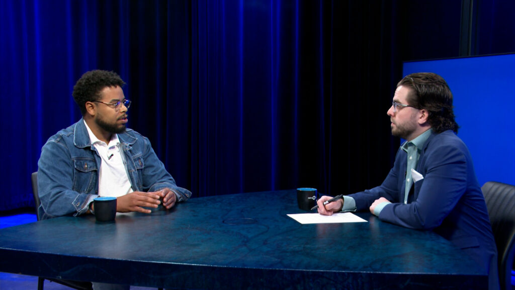 Two men sit across from each other at a blue table in a studio setting, having a conversation. One wears a denim jacket, the other a suit, with papers and mugs on the table.