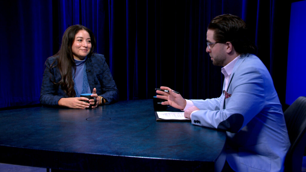 Two people sit across from each other at a round table, engaged in conversation. One holds a mug, the other holds a pen and notepad. The background is dark with blue lighting.