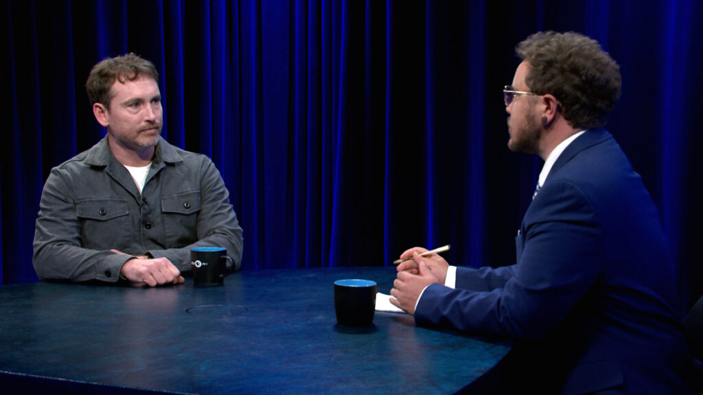 Two men sit across from each other at a round table with mugs, having a discussion in a studio with dark blue curtains.