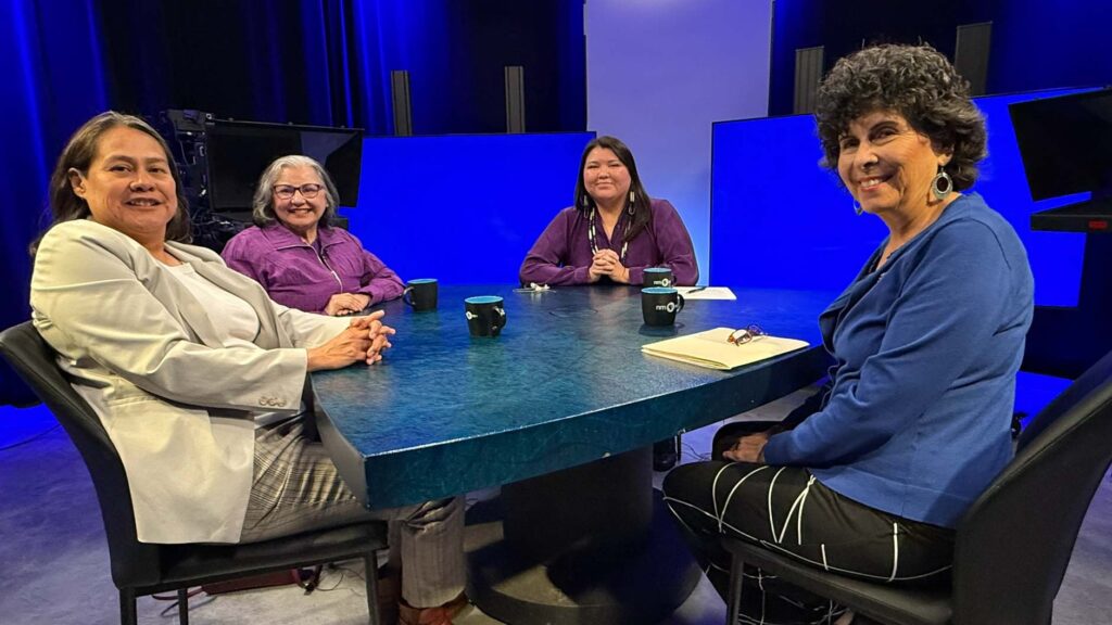 Four women sit around a blue table in a studio, facing the camera, with notebooks and coffee cups in front of them under bright studio lights.