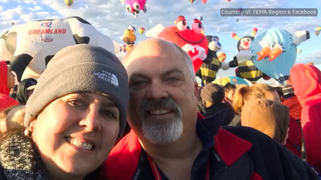 Two people smile for a selfie at an outdoor event with colorful hot air balloons, including ones shaped like bees, floating in the sky behind them.