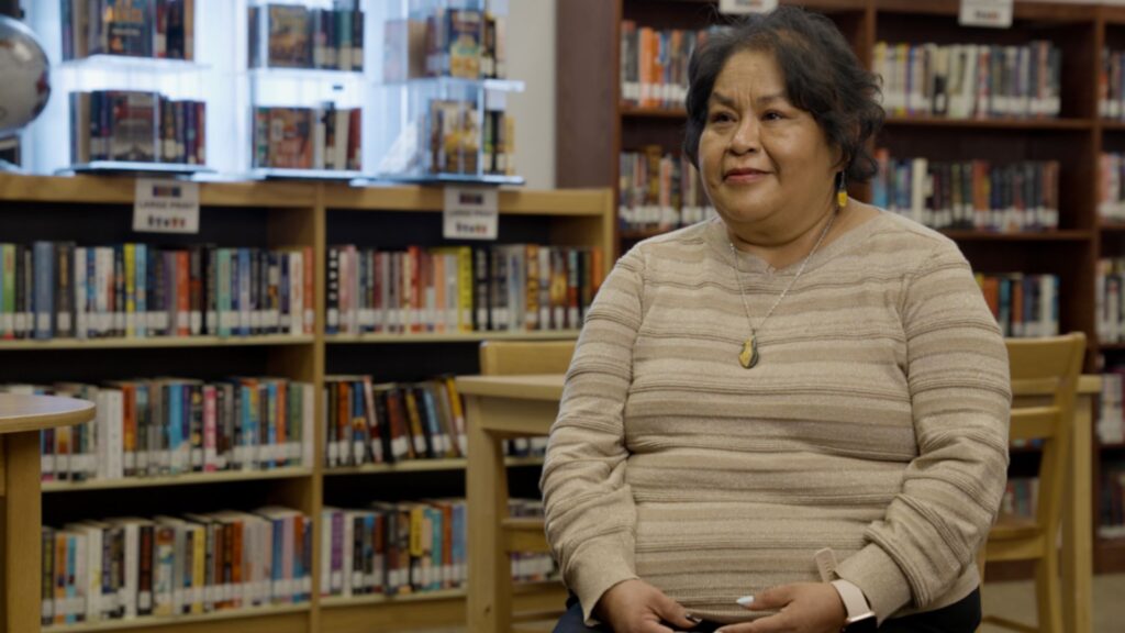 A woman sits in a library, holding a smartphone, with bookshelves filled with books in the background.