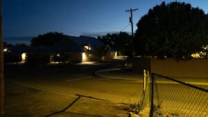 Residential street at dusk with houses lit by exterior lights, a chain-link fence in the foreground, and trees and utility poles under a darkening sky.