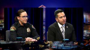 Two people sit at a table during a televised interview; one is a police officer in uniform, and the other is a man in a suit and tie. Cityscape graphics are visible in the background.