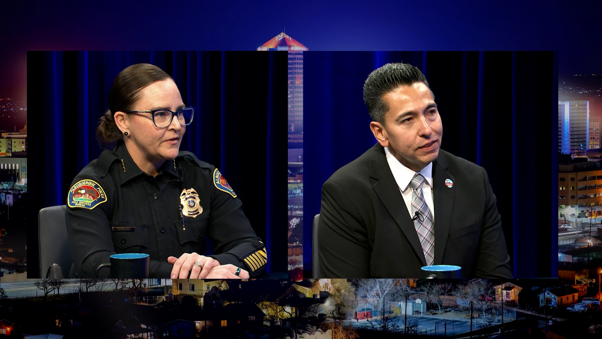 Two people sit at a table during a televised interview; one is a police officer in uniform, and the other is a man in a suit and tie. Cityscape graphics are visible in the background.