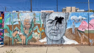 A mural on a concrete wall features a large grayscale portrait with black paint splattered over the face, surrounded by colorful scenes including corn plants and figures.