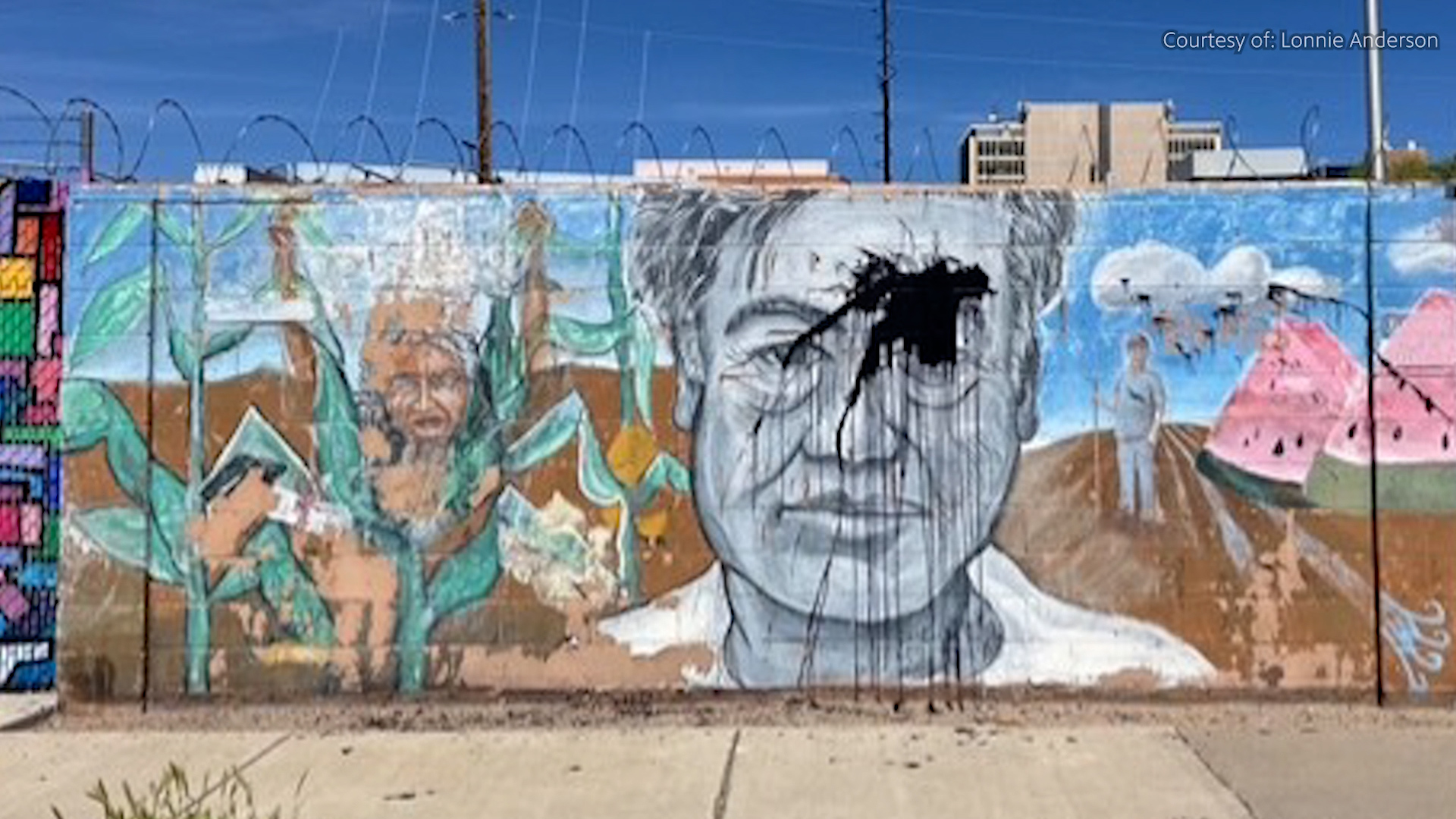 A mural on a concrete wall features a large grayscale portrait with black paint splattered over the face, surrounded by colorful scenes including corn plants and figures.