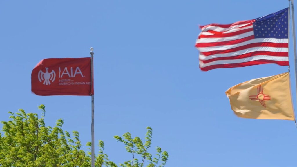 Three flags flying against a blue sky: the IAIA flag, the United States flag, and the New Mexico state flag, with green tree branches visible at the bottom.